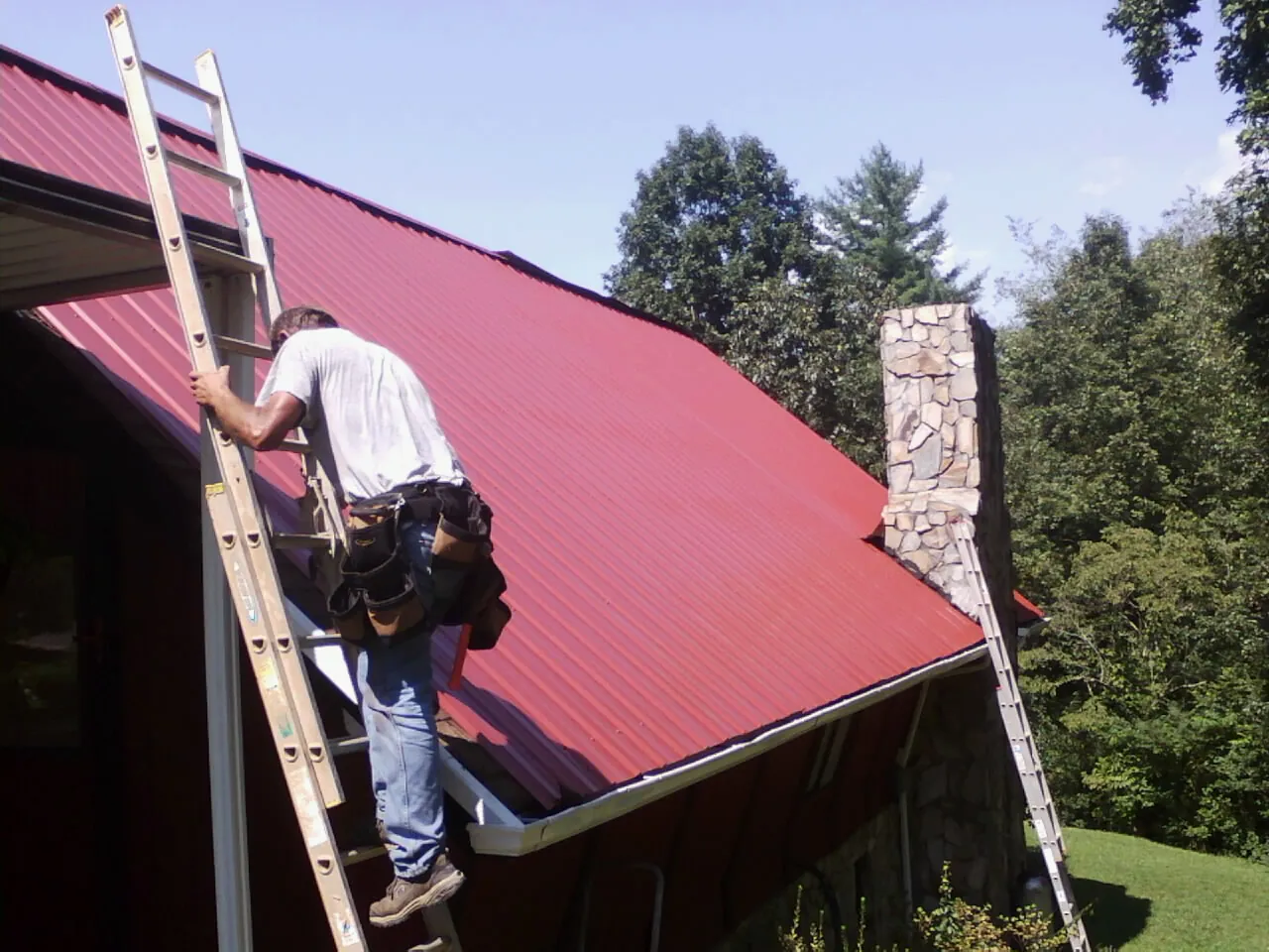 Roofer on a ladder beside a red metal roof and stone chimney — Soffit & Fascia Repair by Roofing Brothers in Stone Park
